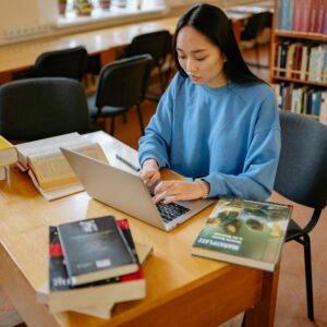 Asian woman typing on a laptop surrounded by books in a library setting.