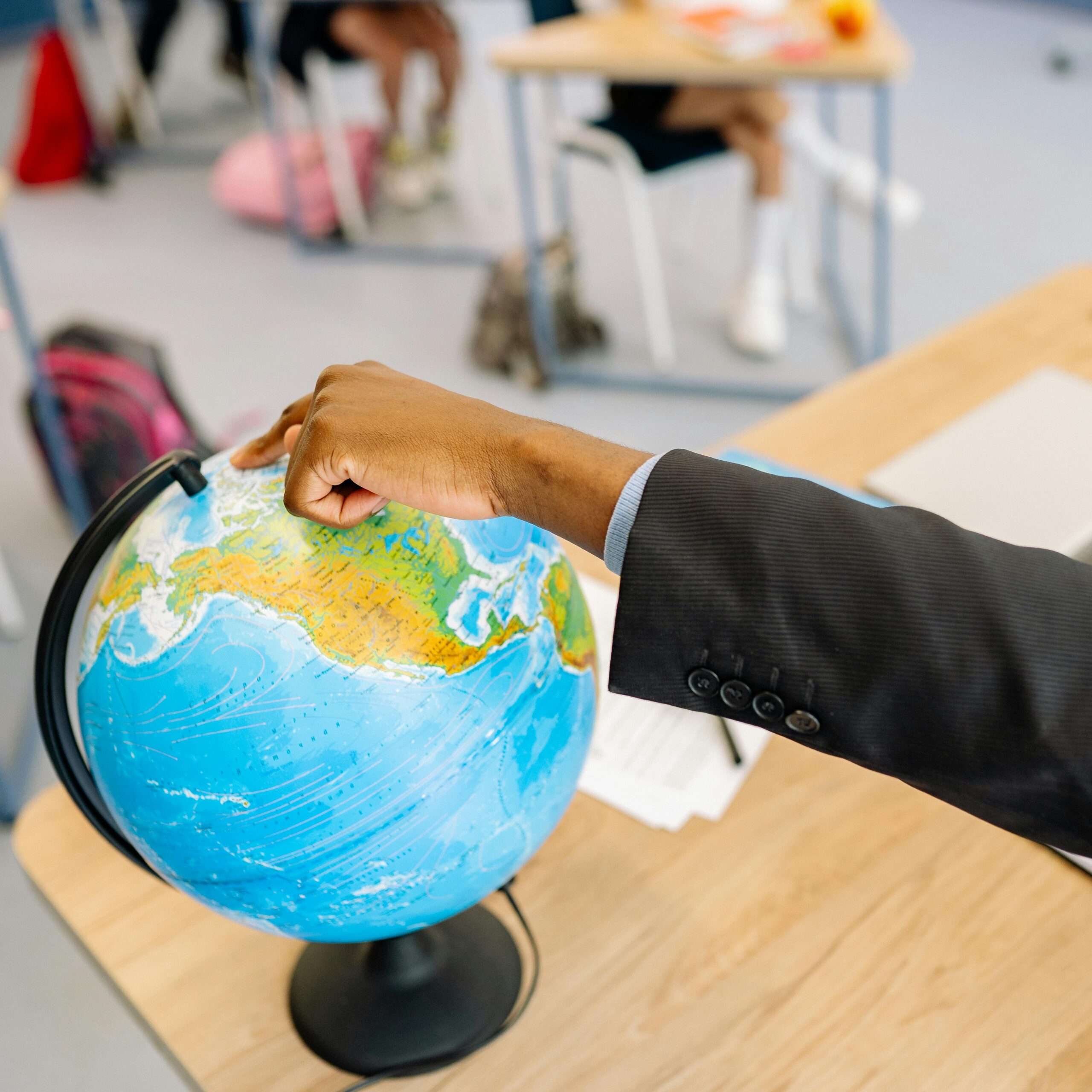 A teacher's hand points at a globe in a classroom while students are present.